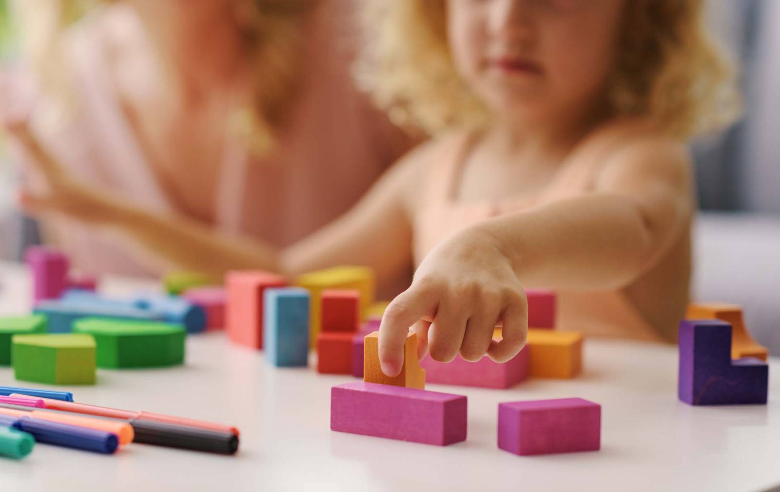 A toddler stacking wooden blocks at a table while an adult guides her hand